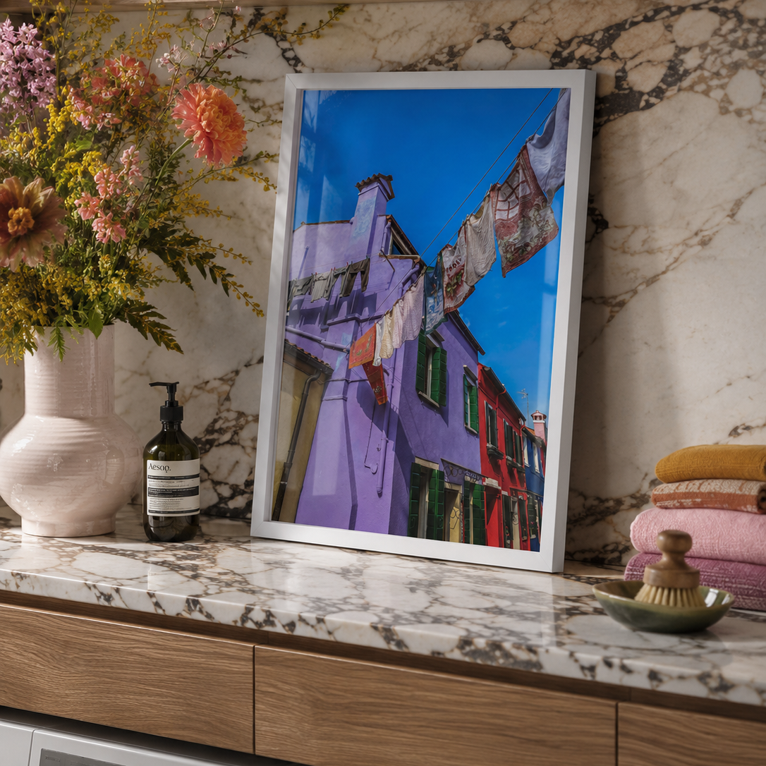 A framed art print depicting colorful purple and red buildings under a bright blue sky, styled on a marble countertop in a modern laundry room setting.