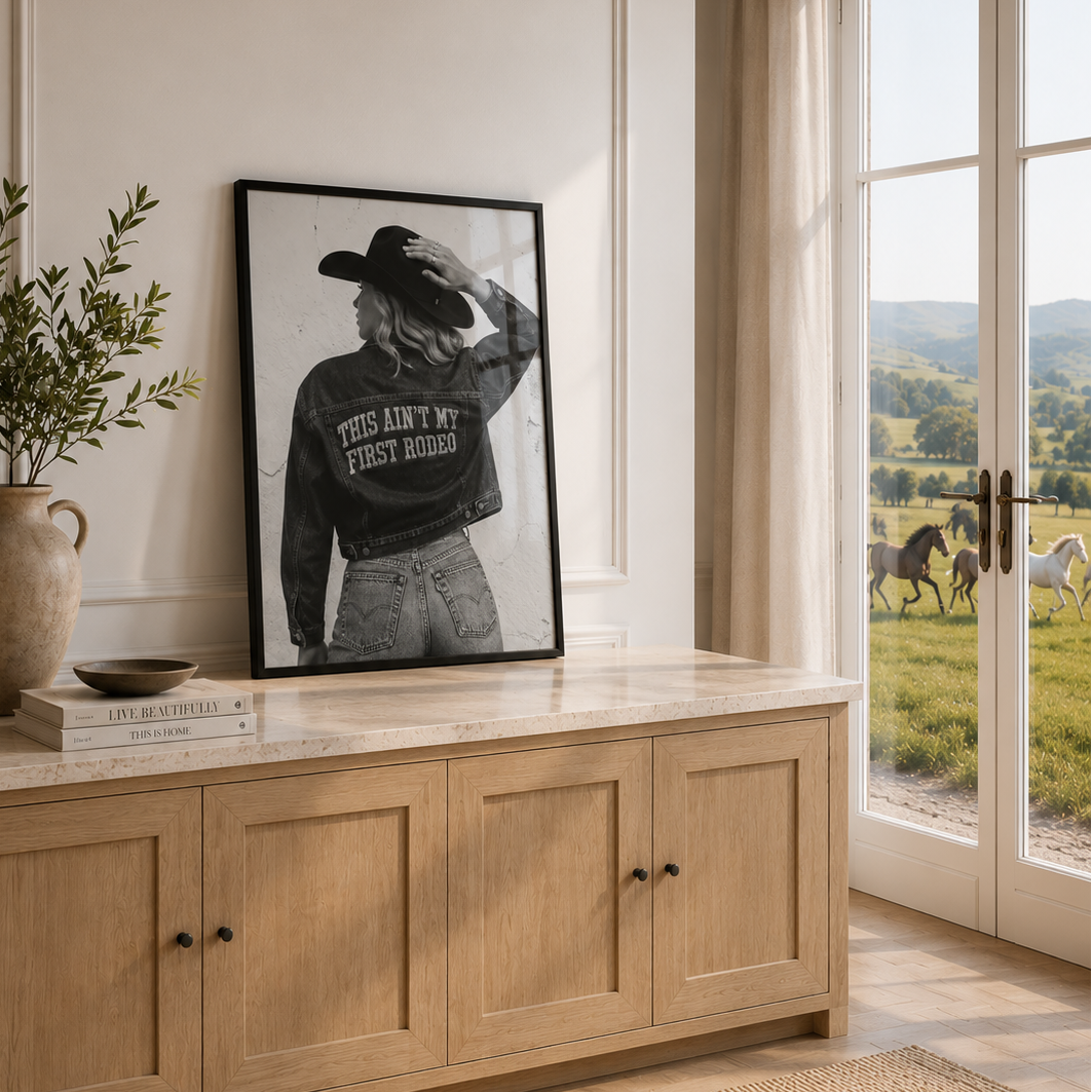 A framed black and white western-style poster leaning on a light wood console cabinet in a bright room, with a window showing a horse pasture outside.