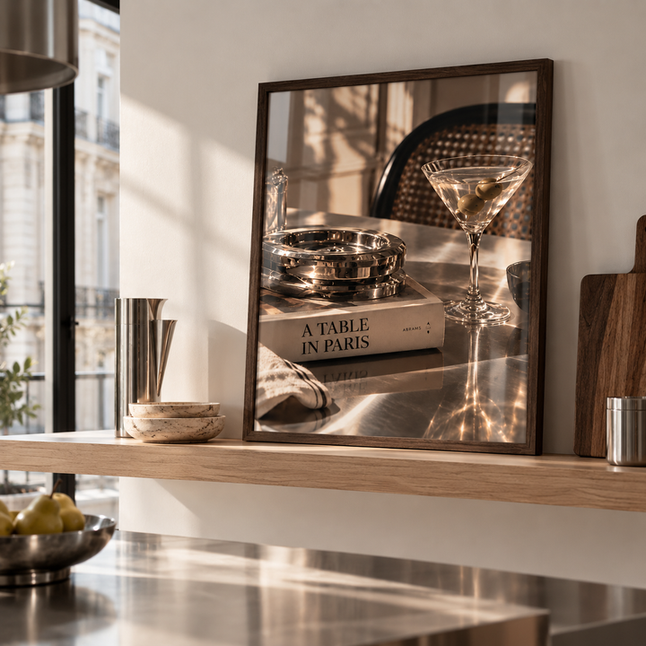 A framed art print with a dark wood frame is displayed on a light wood floating shelf in a modern kitchen setting. The print features a warm-toned photograph of a martini glass, a metal ashtray, and a book titled 'A TABLE IN PARIS'.