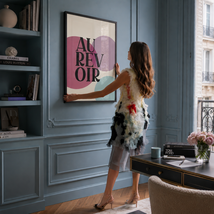A woman stands in a blue paneled room inspired by Emily in Paris, holding a framed poster that reads 'AU REVOIR' with pink, cream, and black shapes. The room includes built-in shelves with books, a desk with flowers, and a window overlooking a building exterior.