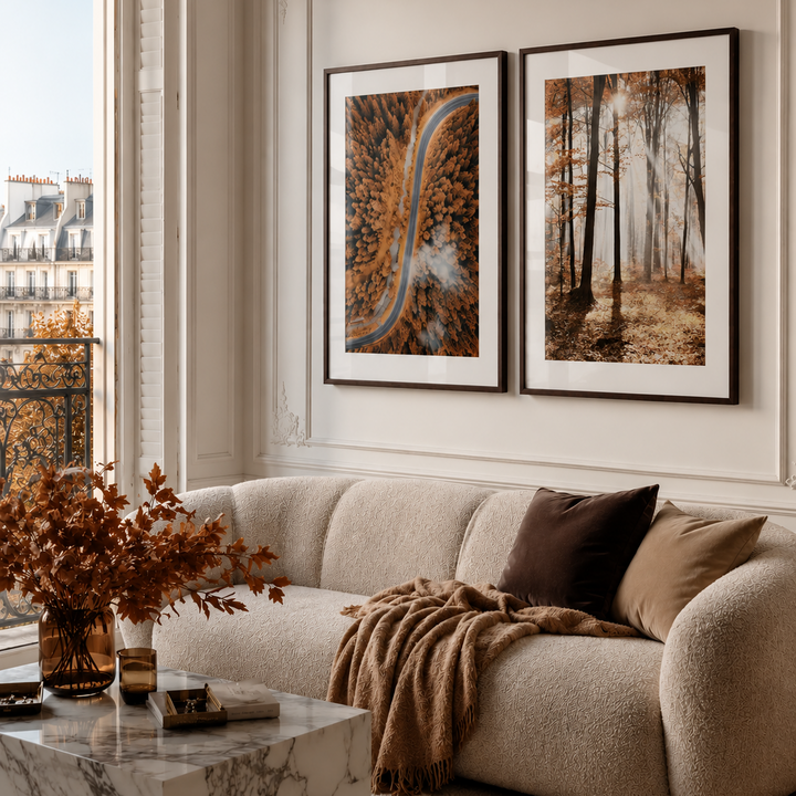 A styled living room interior with a cream boucle sofa, brown throw blanket, marble coffee table, and two framed wall art prints hanging above the couch. The prints depict abstract and forest scenes in warm autumn tones.