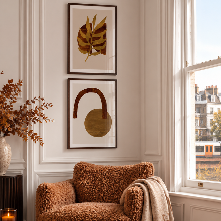 A styled interior corner with a textured rust-brown armchair beside a window, a cream throw blanket draped over the arm, and two framed abstract botanical and geometric prints on the wall.