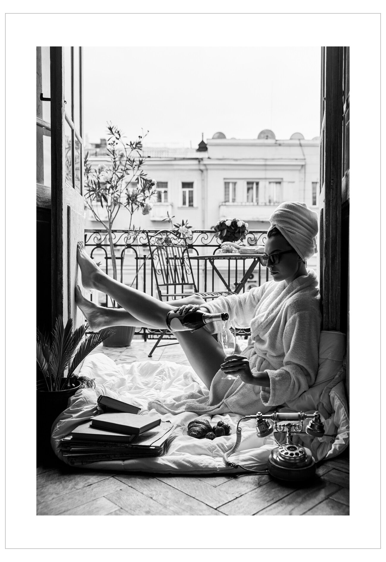 Person in a bathrobe sitting on a balcony with books and a plant, wearing a towel on their head.