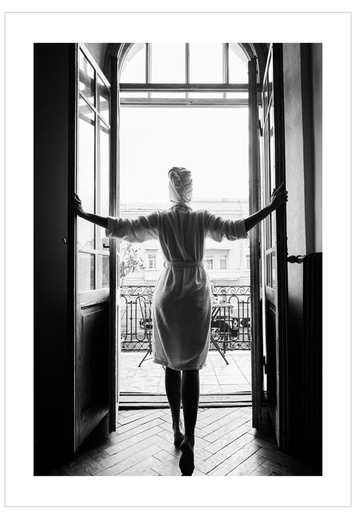 Person in a white coat standing in a doorway with arms outstretched, looking out onto a balcony.