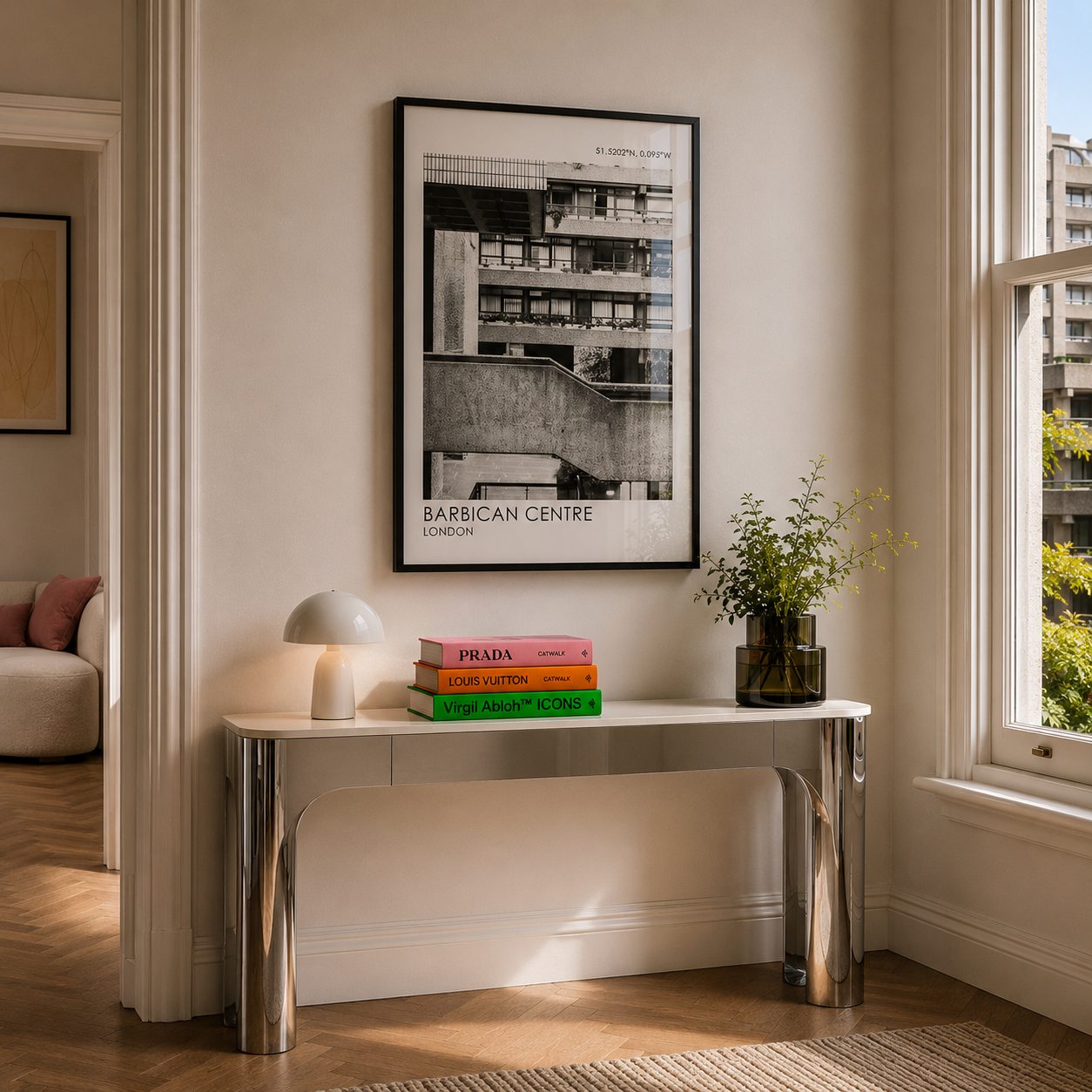 Modern interior with a table holding Prada books, a lamp, and a vase with plants, framed artwork on the wall.