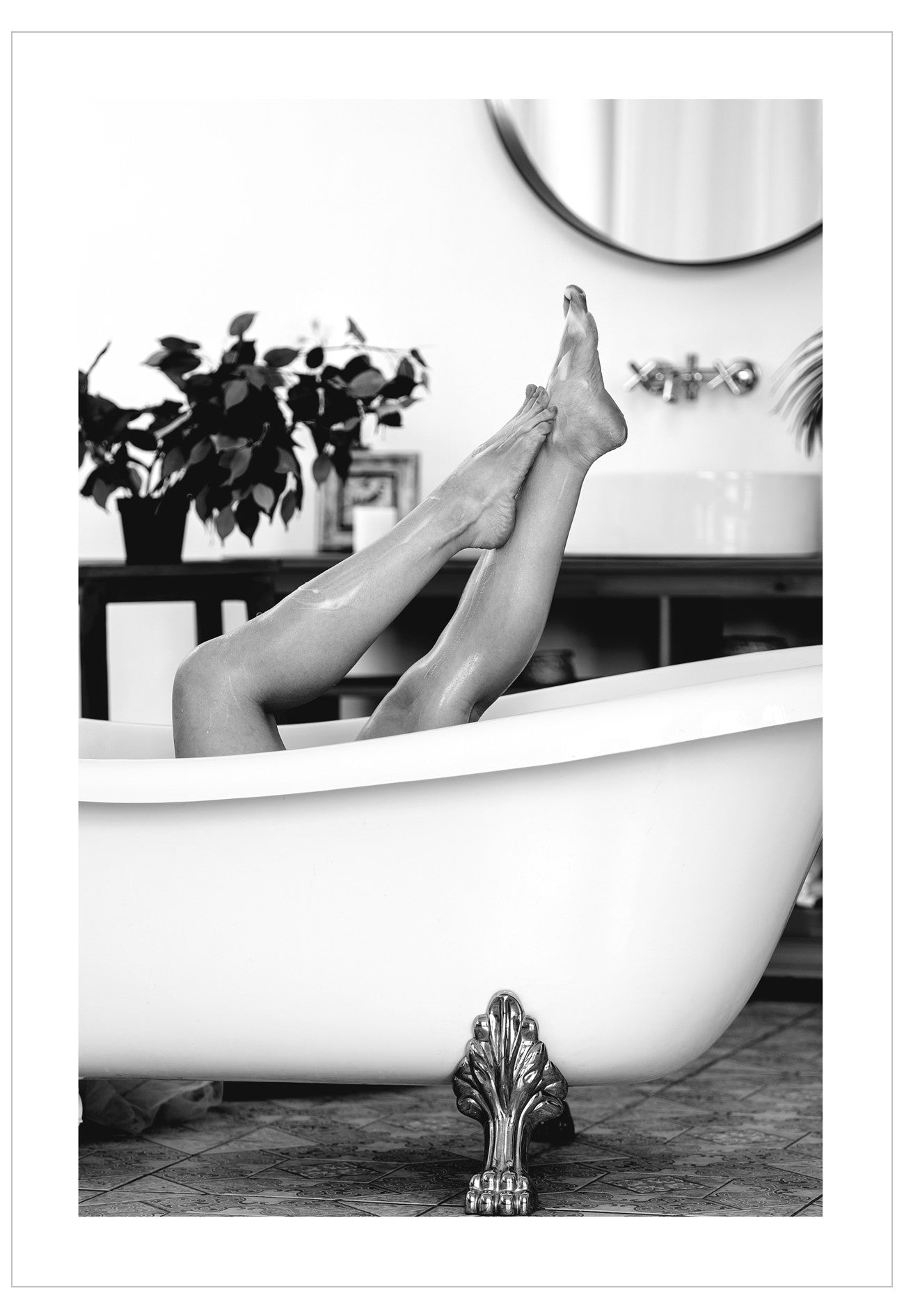 Person's legs in a bathtub with a decorative clawfoot design, surrounded by plants and a mirror.
