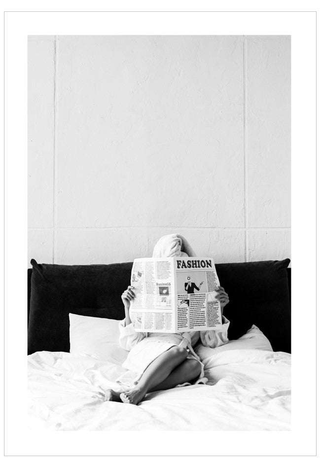 Person sitting on a bed holding a newspaper with a fashion section, wearing a headscarf.