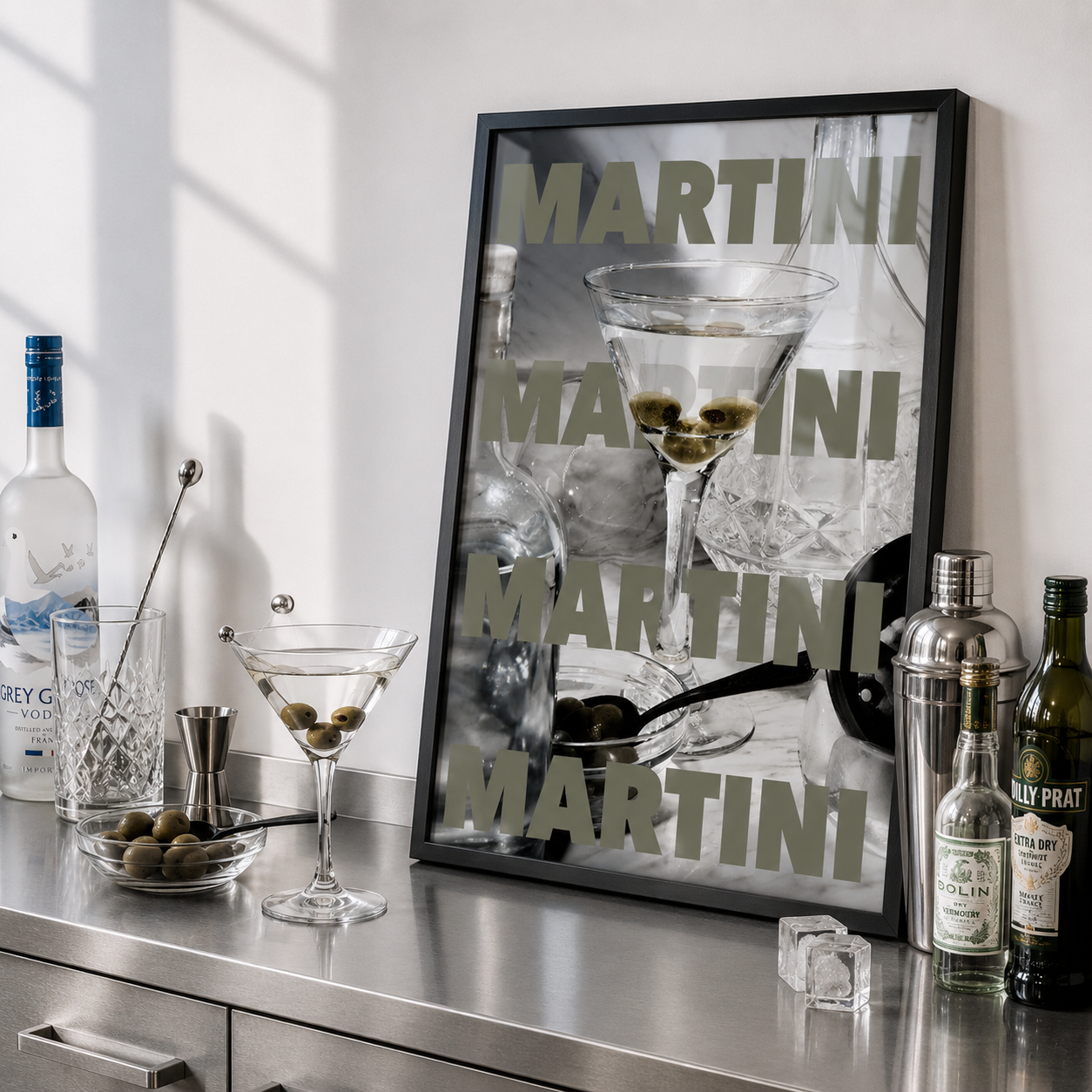 Bar setup with a framed martini glass poster, bottles, and glasses on a metallic surface.