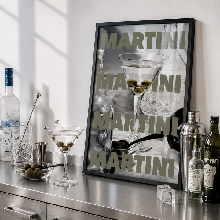Bar setup with a framed martini glass poster, bottles, and glasses on a metallic surface.