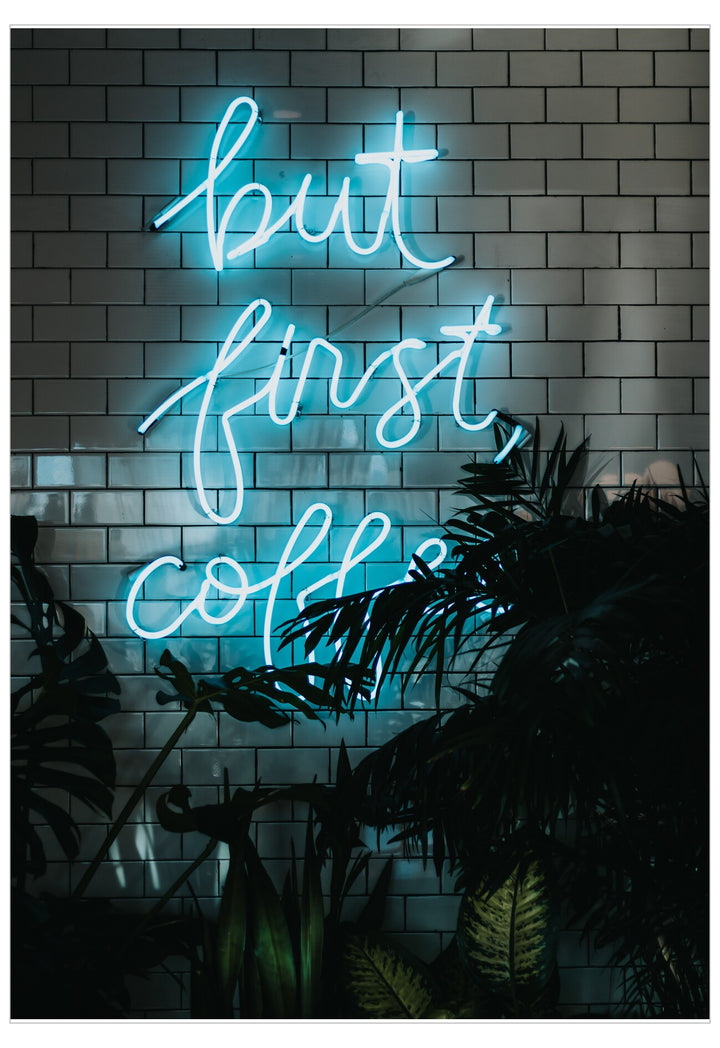 Neon sign with 'but first, coffee' text on a brick wall with plants.