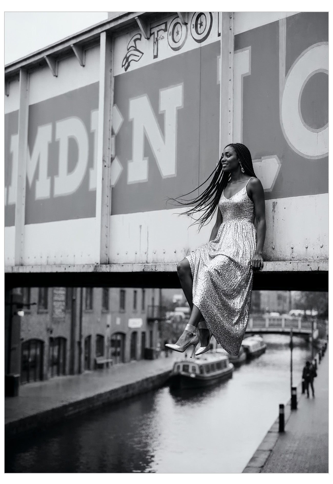 Woman in a long dress sitting on Camden bridge over a canal with a large sign in the background.