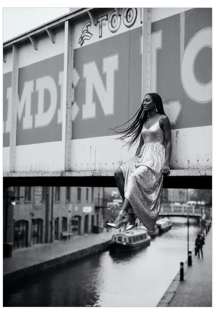 Woman in a long dress sitting on Camden bridge over a canal with a large sign in the background.