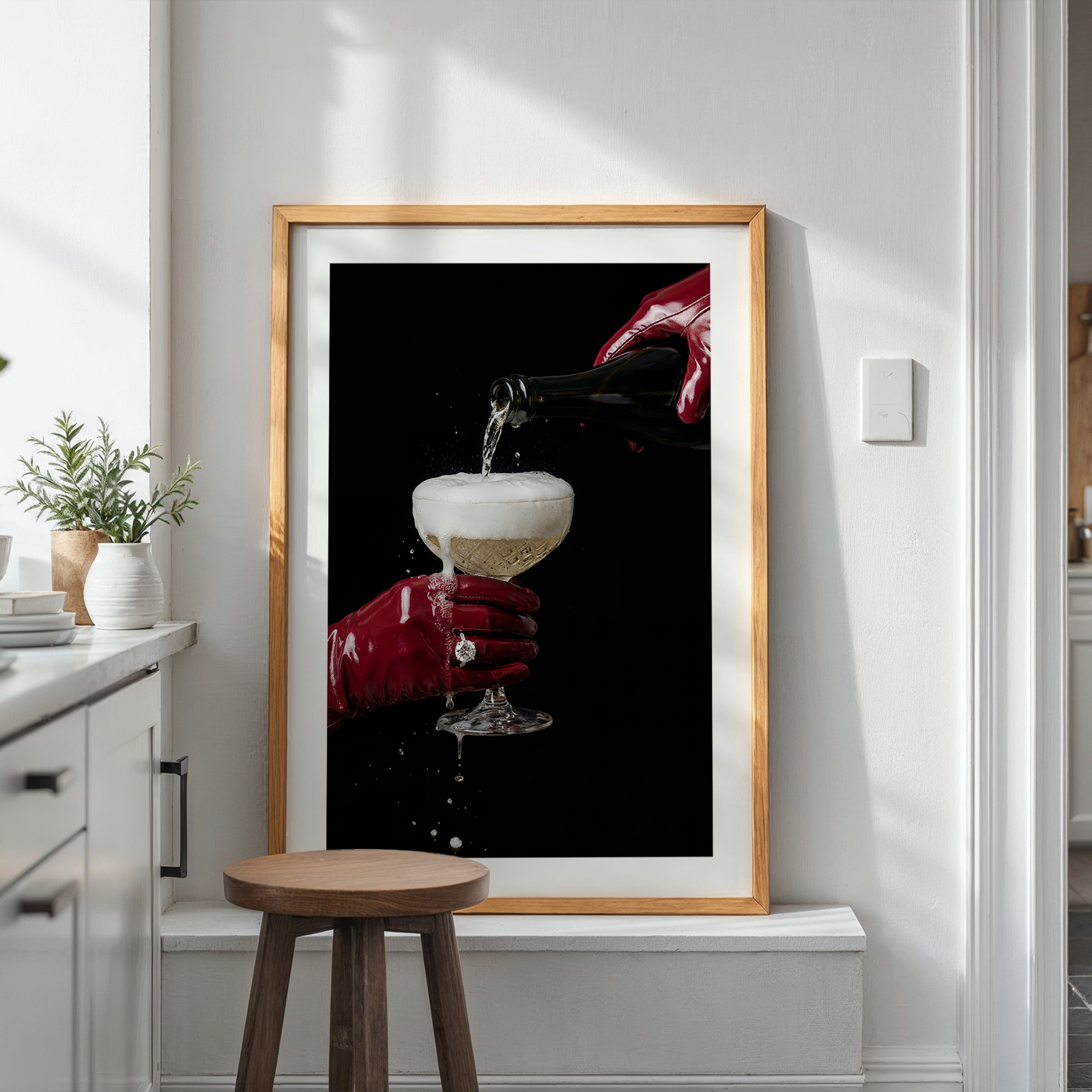 Framed artwork of a hand pouring a champagne drink into a glass on a white wall with plants and a stool below.