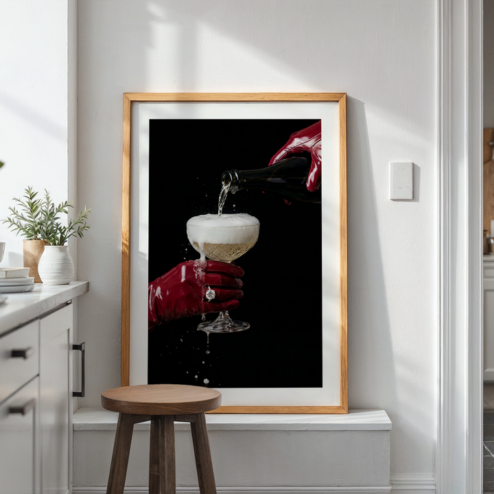 Framed artwork of a hand pouring a champagne drink into a glass on a white wall with plants and a stool below.