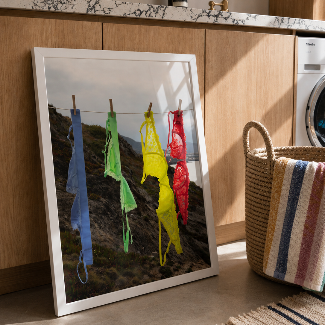 A framed art print leaning against a wooden laundry room cabinet. The print shows colorful bikini-style garments hanging on a clothesline against a mountain background.