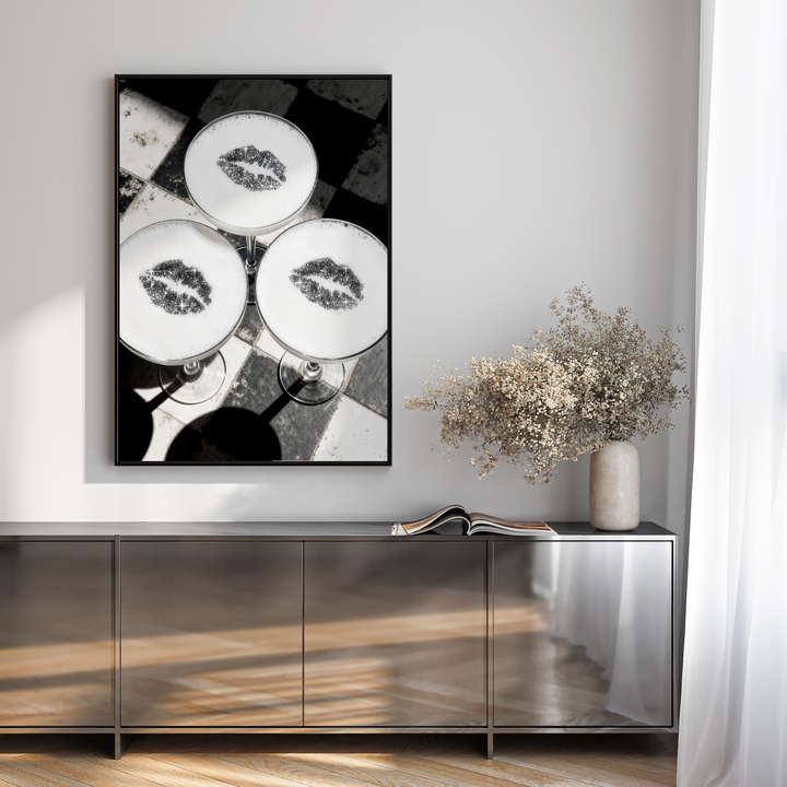 Framed artwork on a wall above a reflective surface with a vase and flowers.