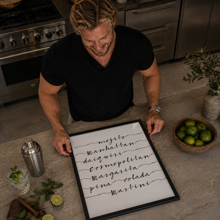 Man in a kitchen holding a framed list of cocktail names with limes and a cocktail shaker on the counter.