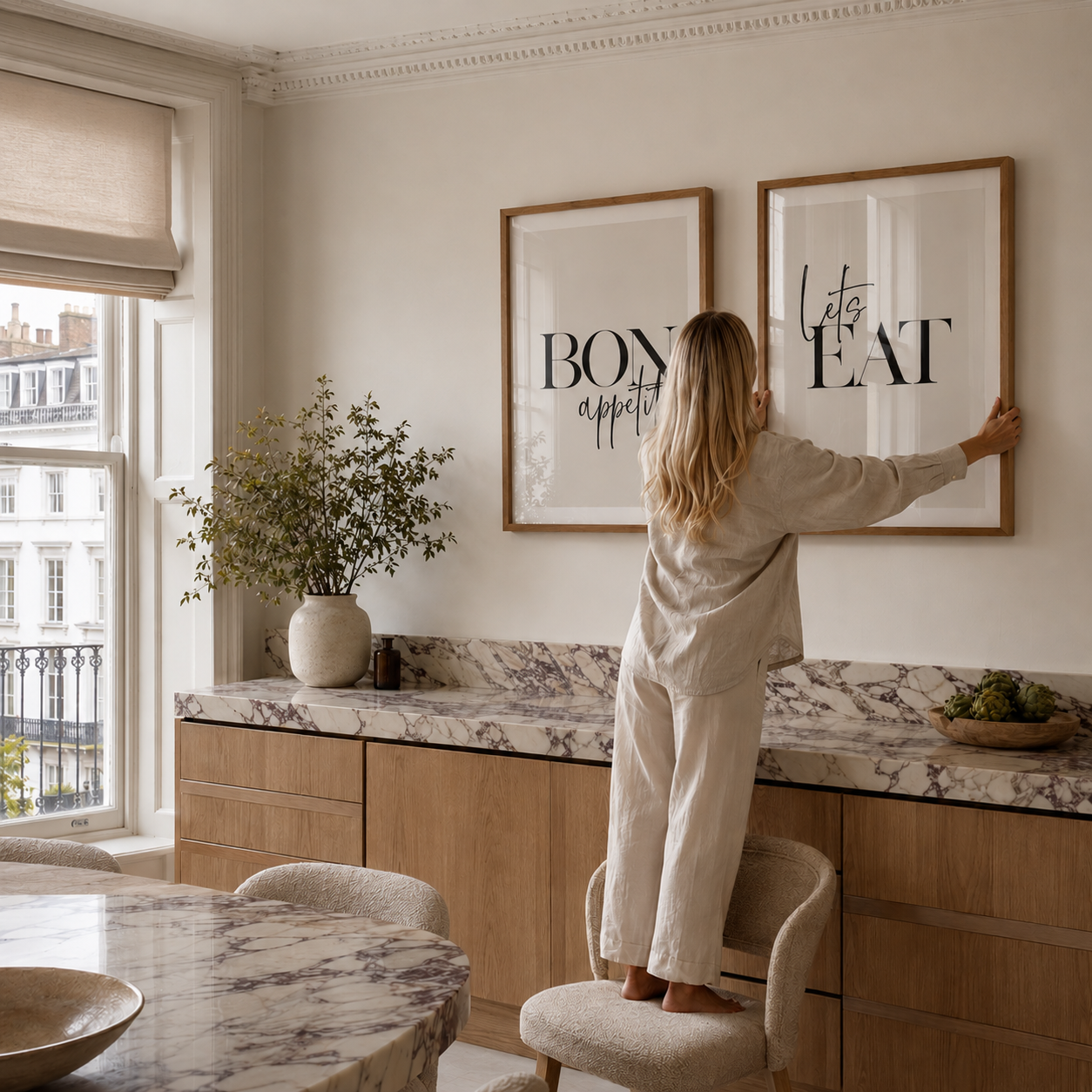 Person hanging a framed picture in a stylish kitchen with marble countertops and large windows.