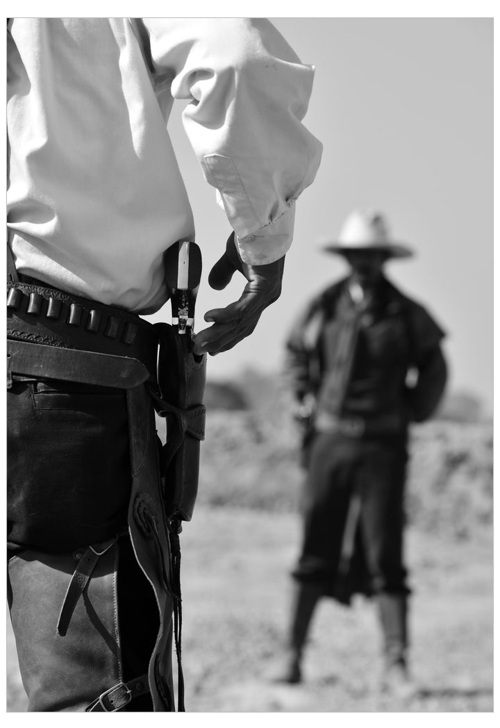 Black and white photograph of a cowboy in a western outfit with a holster at the waist, with another cowboy standing blurred in the background outdoors.