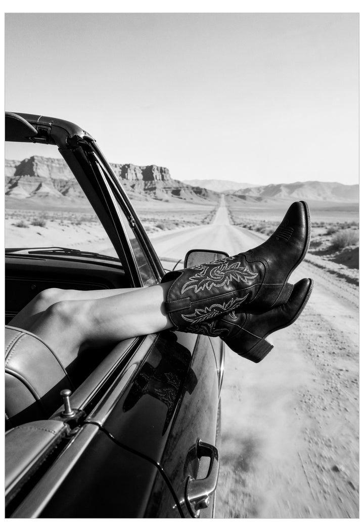 Black and white photograph of a person's legs stretched out of a convertible car on a desert road, wearing black western cowboy boots with intricate embroidery.