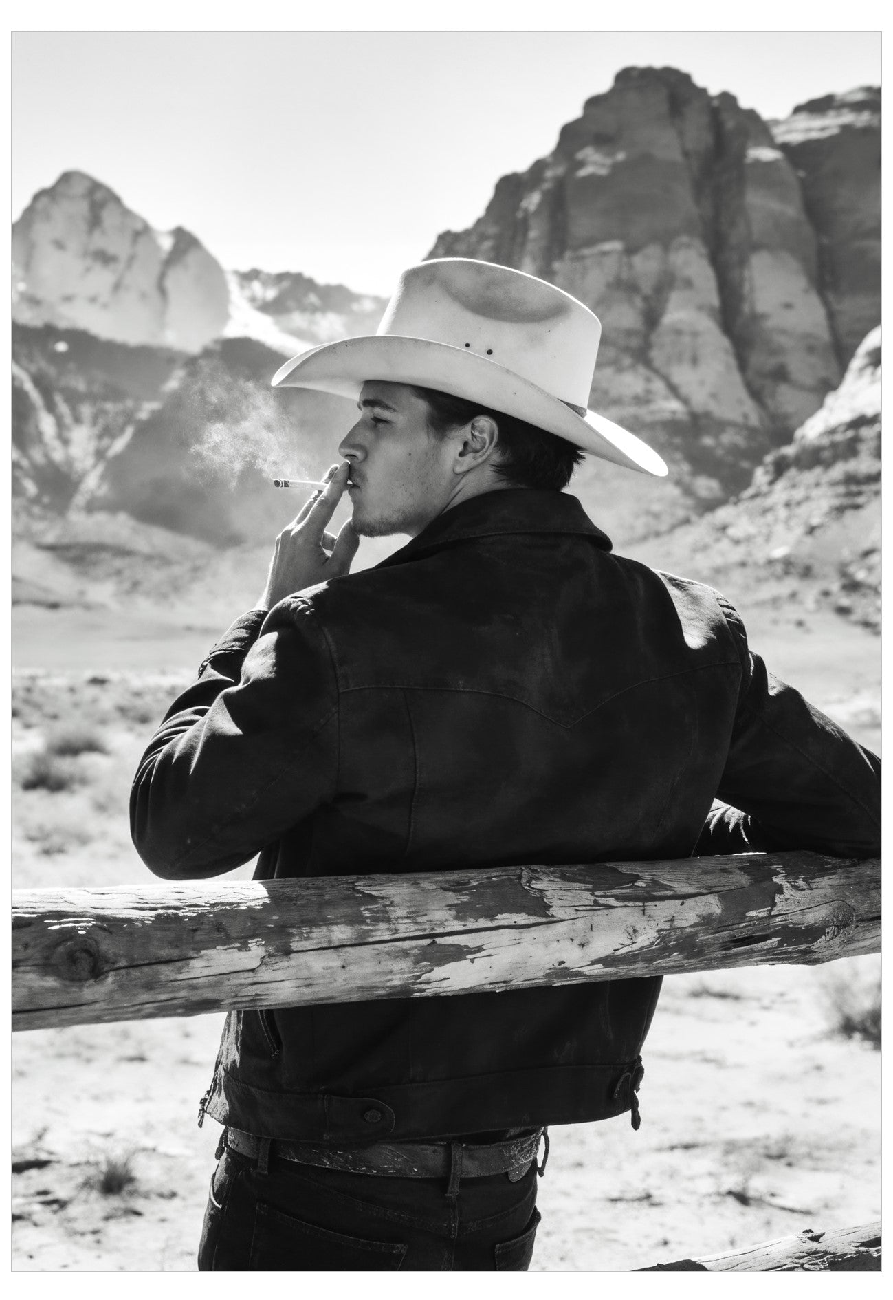 A black and white photograph of a man in a cowboy hat and dark jacket, leaning on a wooden fence in a desert landscape with mountains in the background.