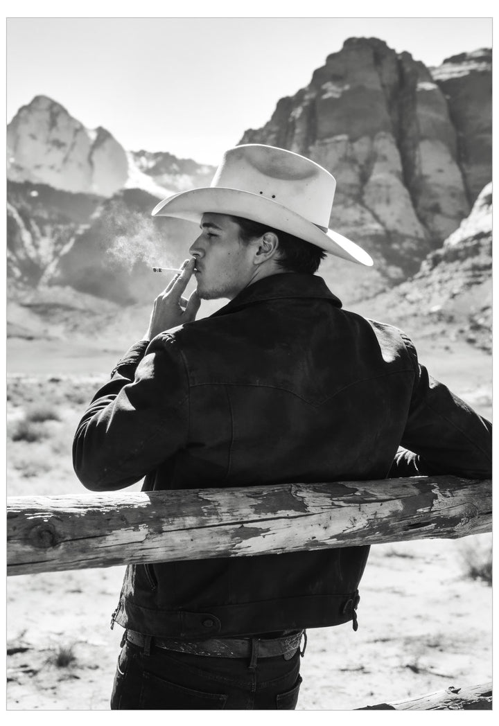 A black and white photograph of a man in a cowboy hat and dark jacket, leaning on a wooden fence in a desert landscape with mountains in the background.