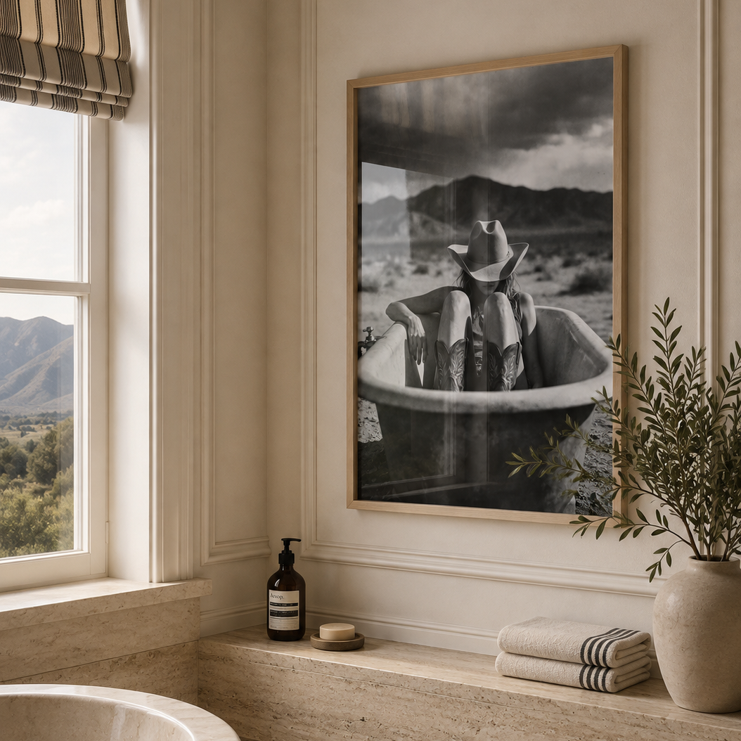 Black and white photography poster of a cowgirl in a desert bath tub in a large oak frame in a luxury country home bathroom with Travertine finishes.