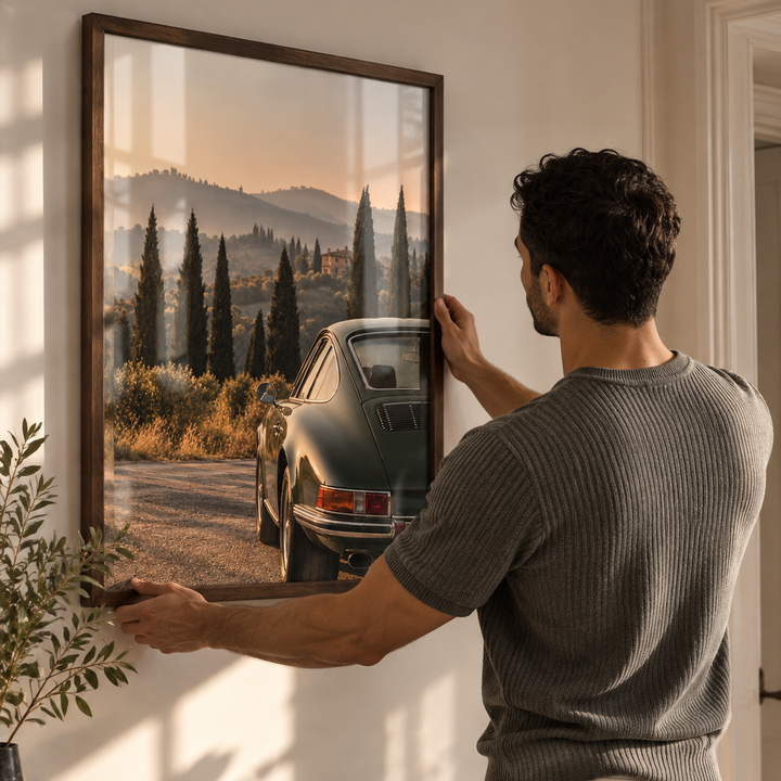 A man in a ribbed gray t-shirt and white pants hangs a large framed landscape print on a beige wall above a modern console table. The print depicts a vintage green car on a dirt road surrounded by cypress trees at sunset.
