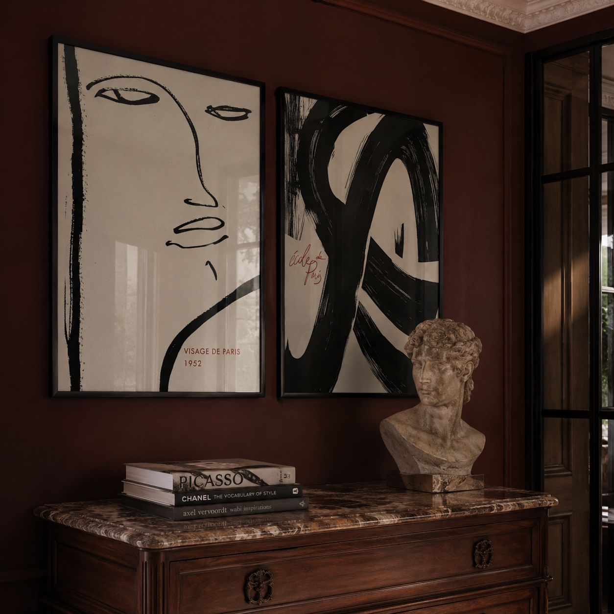 Two abstract artworks on a wall above a wooden cabinet with books and a bust.
