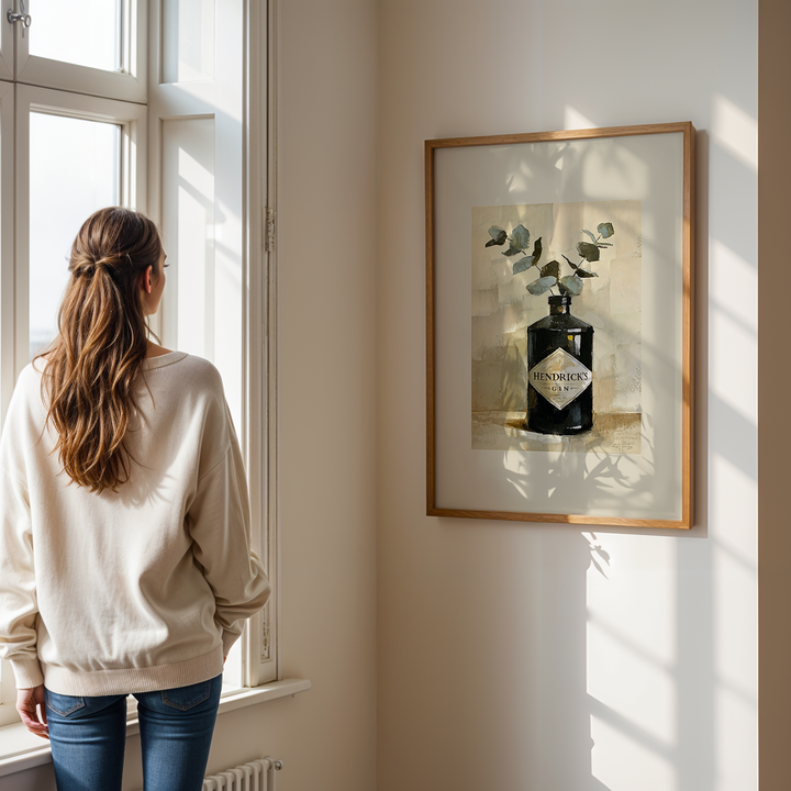 Person standing by a window looking out, with a framed picture of a bottle on the wall.