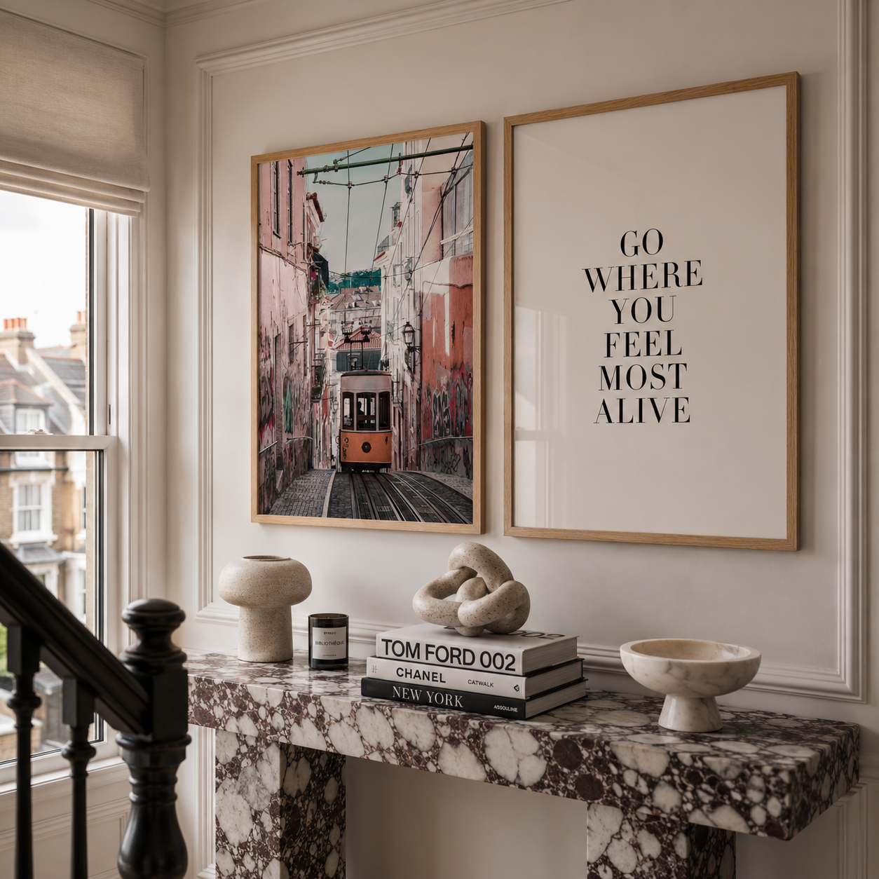 Two framed wall art prints displayed above a marble console table in a styled interior. The left print shows a street scene with a tram in a narrow city street, and the right print features the text 'GO WHERE YOU FEEL MOST ALIVE' in black on a white background.