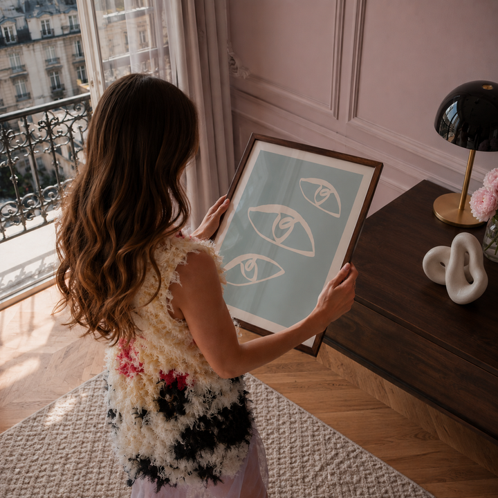 A woman in a textured Chanel cream and black dress holds a framed art print featuring three minimalist white eyes on a light blue background, set in a stylish room inspired by Emily in Paris with a dark wood table and a window.