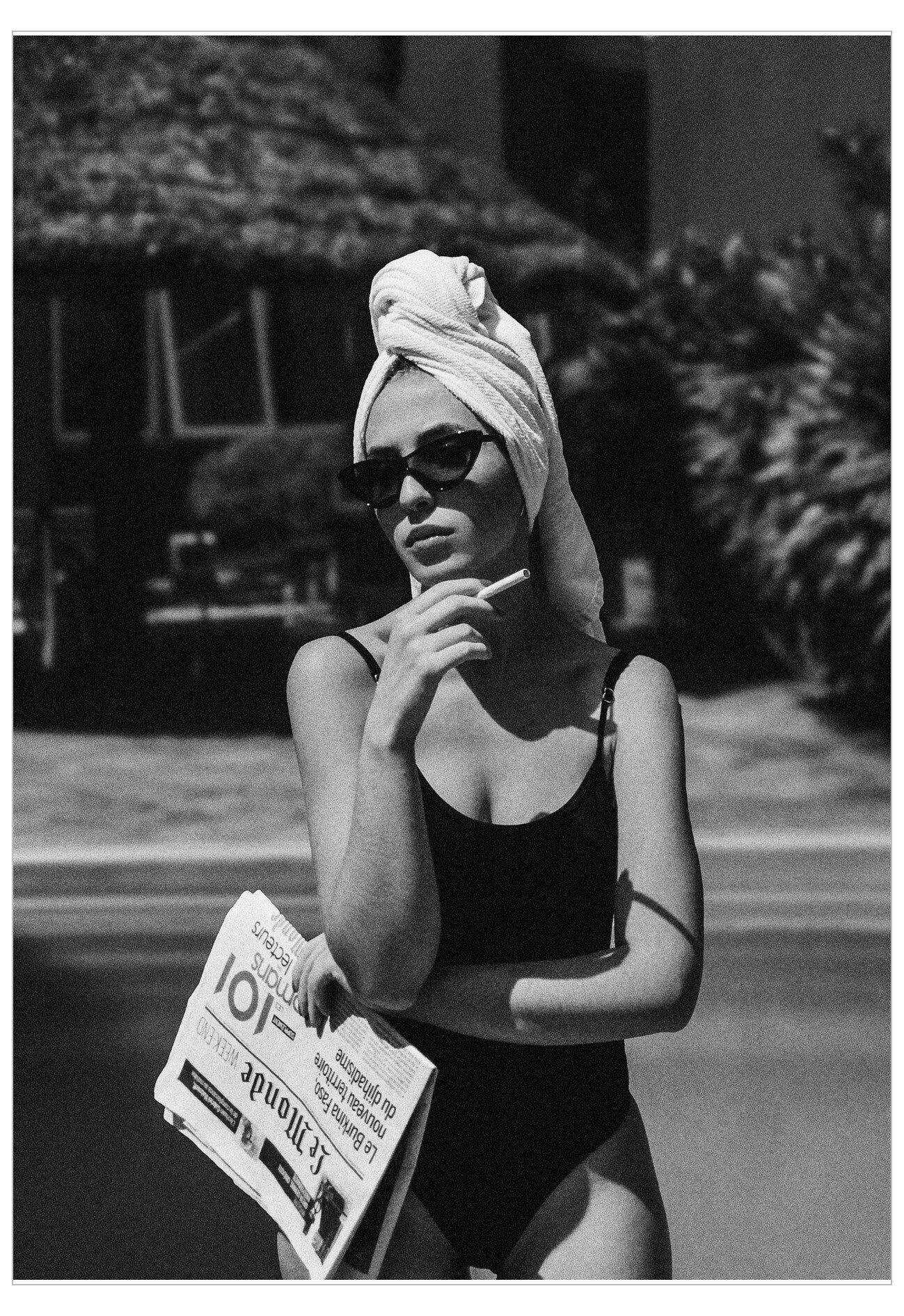 Woman in a black swimsuit with a towel on her head, holding a newspaper by a pool.