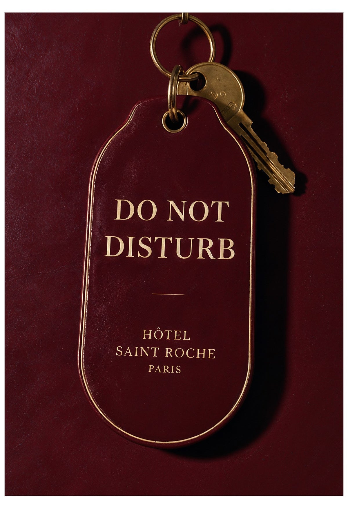 A dark burgundy hotel key tag with gold lettering reading 'DO NOT DISTURB' and 'HÔTEL SAINT ROCHE PARIS', attached to a gold key ring with a brass key, shown against a matching burgundy background.