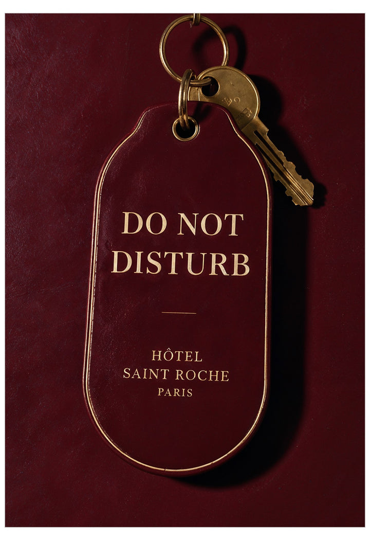A dark burgundy hotel key tag with gold lettering reading 'DO NOT DISTURB' and 'HÔTEL SAINT ROCHE PARIS', attached to a gold key ring with a brass key, shown against a matching burgundy background.