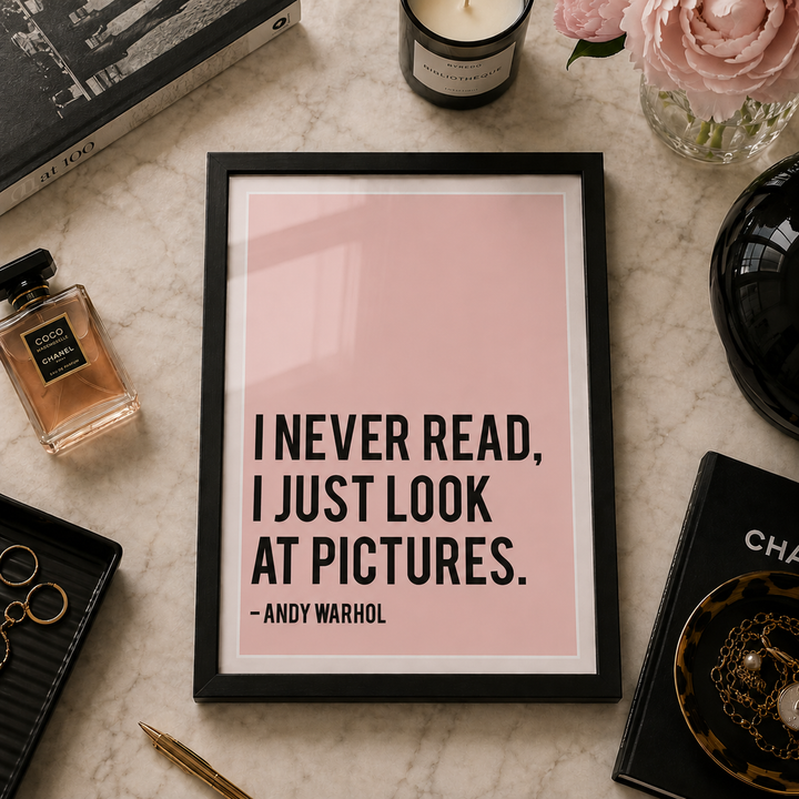 A framed quote print featuring the text 'I NEVER READ, I JUST LOOK AT PICTURES. -ANDY WARHOL' in black on a soft pink background, styled in a flat lay with a candle, perfume bottle, rings, and other decorative items on a marble surface.