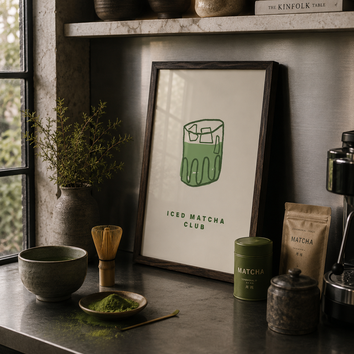 Matcha tea setup with a framed print, bowl, whisk, and containers on a wooden surface.