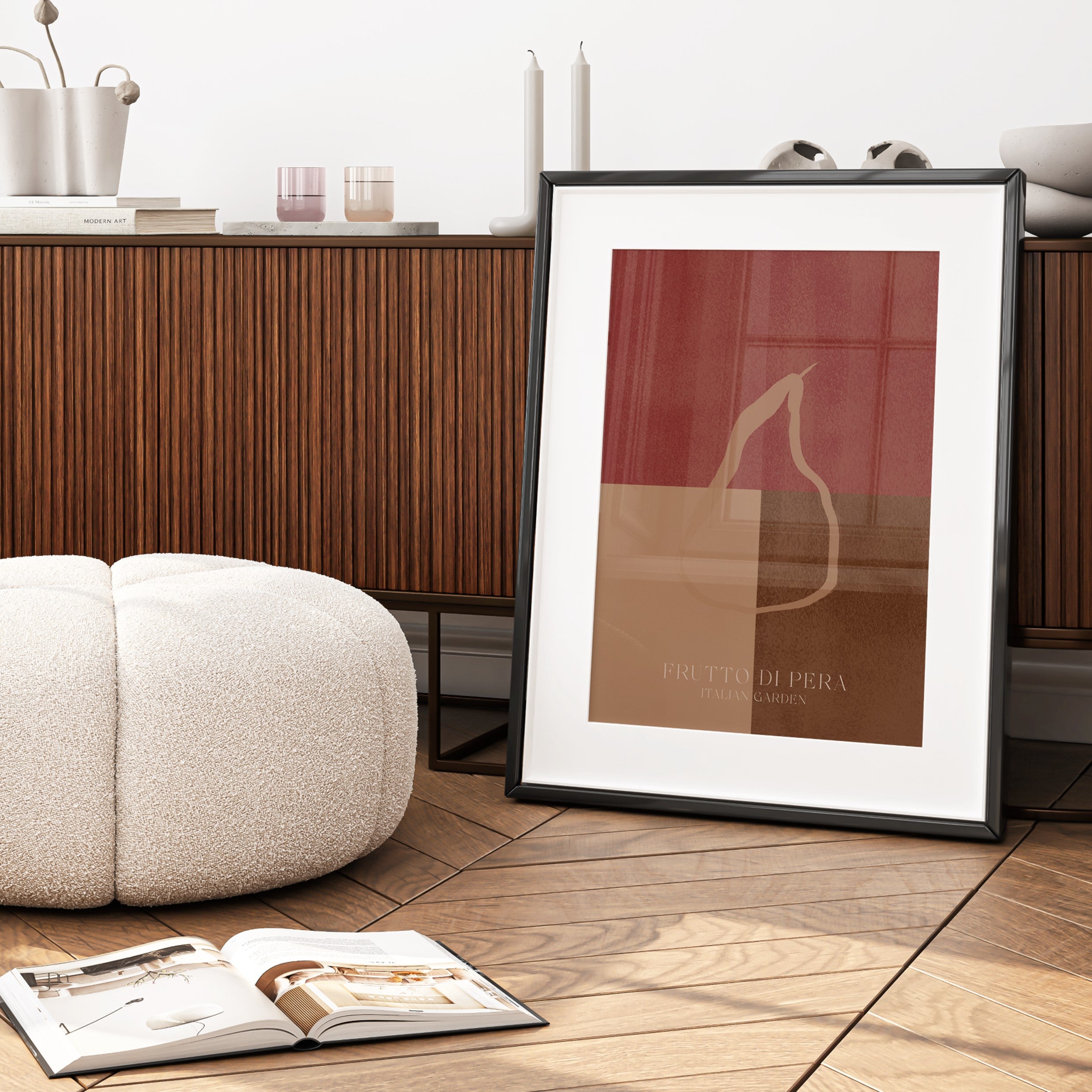 Framed abstract art print on a wooden floor with a white ottoman and open book.