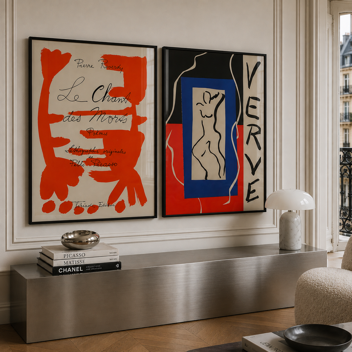 Two framed abstract art prints hanging side-by-side on a cream-colored wall above a modern metallic console table. The left print features bold red abstract shapes on a cream background with black script text, and the right print features black, red, and blue abstract shapes with the word 'VERVE' in black.