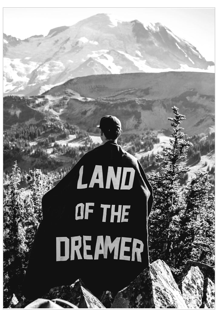 Person wrapped in a blanket with 'LAND OF THE DREAMER' text, standing in a mountainous landscape.