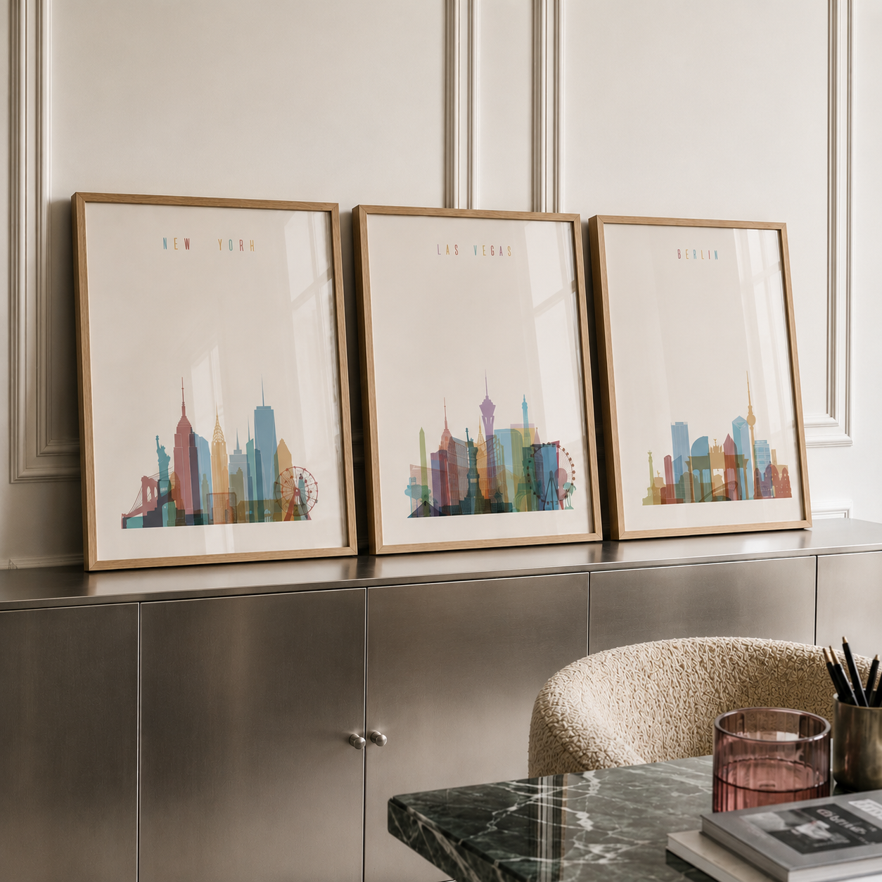Three framed city skyline art prints leaning against a metallic cabinet in a styled interior setting. The prints feature colorful watercolor-style skyscrapers labeled New York, Los Angeles, and Berlin, with light wood frames.