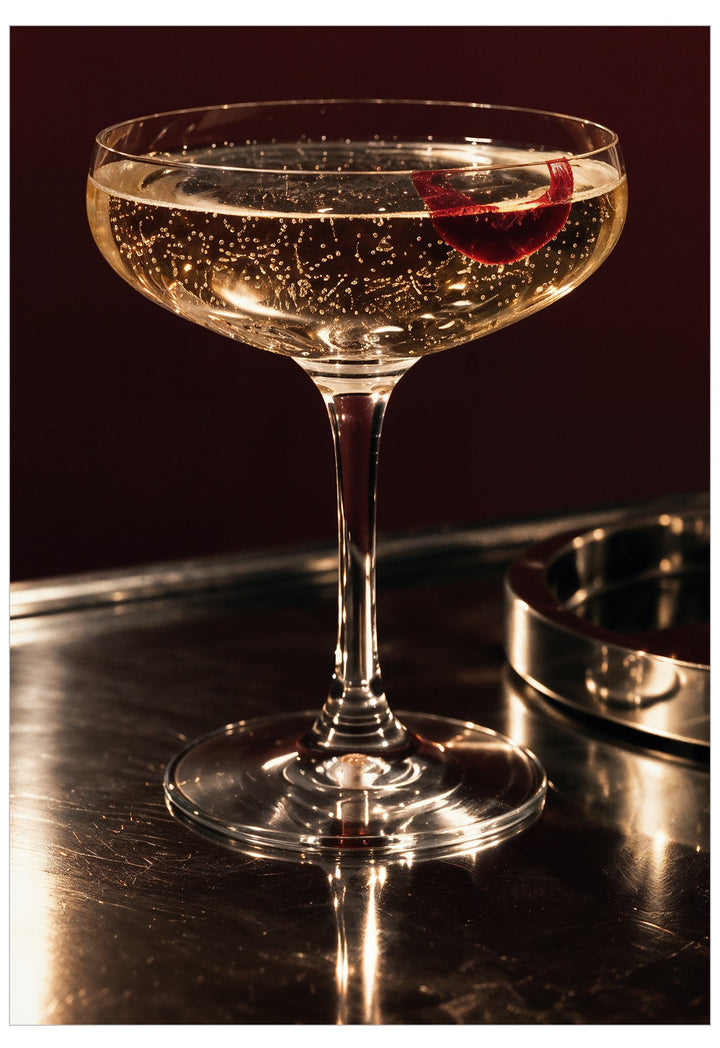 A coupe glass filled with a bubbly golden cocktail, garnished with a red citrus slice, set on a dark bar surface with a metal tray in the background.