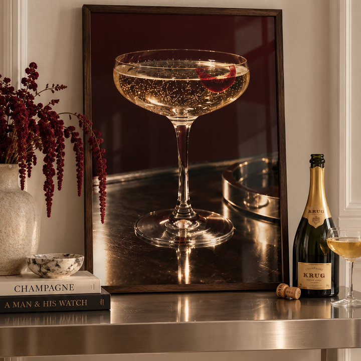 A coupe glass filled with a bubbly golden cocktail, garnished with a red citrus slice, set on a dark bar surface with a metal tray in the background.