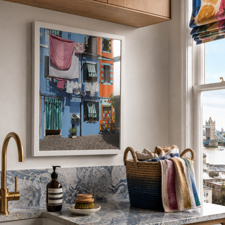 A framed art print depicting a colorful street scene with laundry hanging on lines, styled in a modern laundry room setting above a marble countertop with a Miele washing machine.