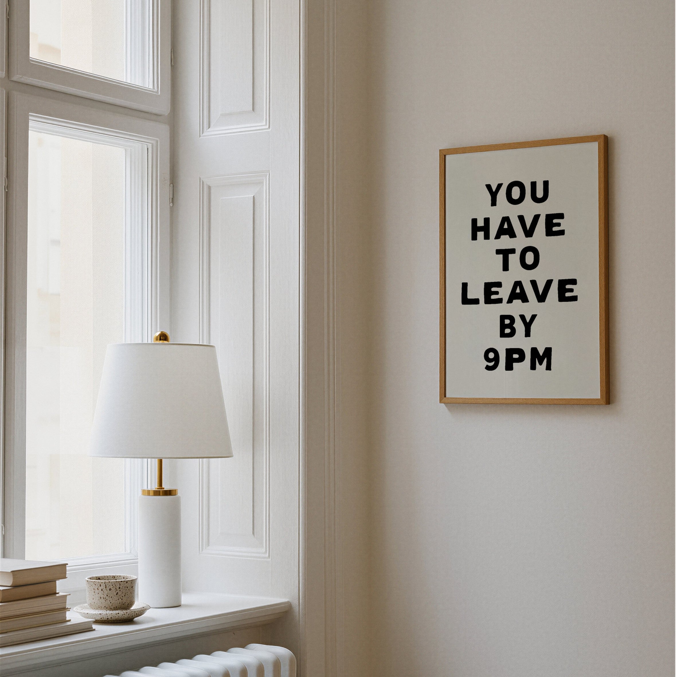 Framed artwork on a wall with a lamp and books on a windowsill.