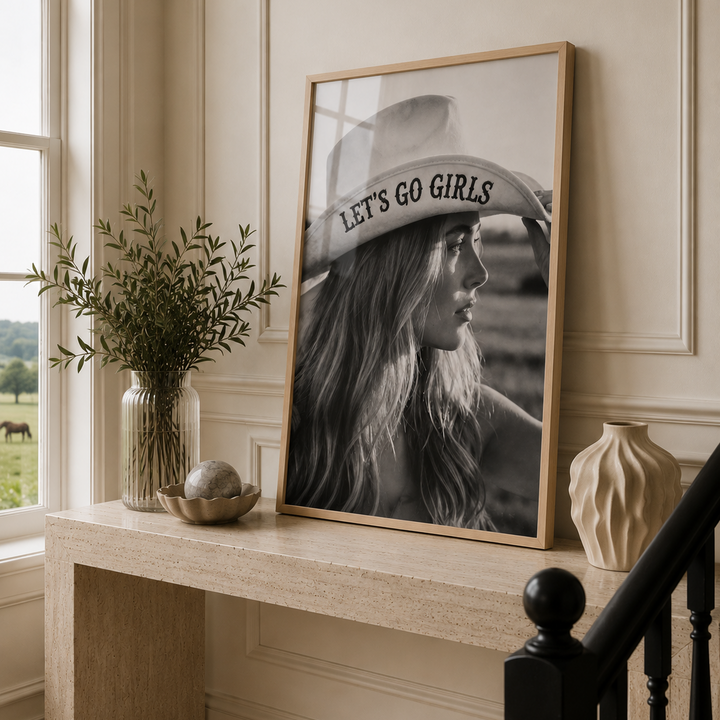 A black and white lifestyle photograph of a woman in profile wearing a cowboy hat with the text 'LET'S GO GIRLS' printed on the brim. In a oak frame in a luxury country hallway.