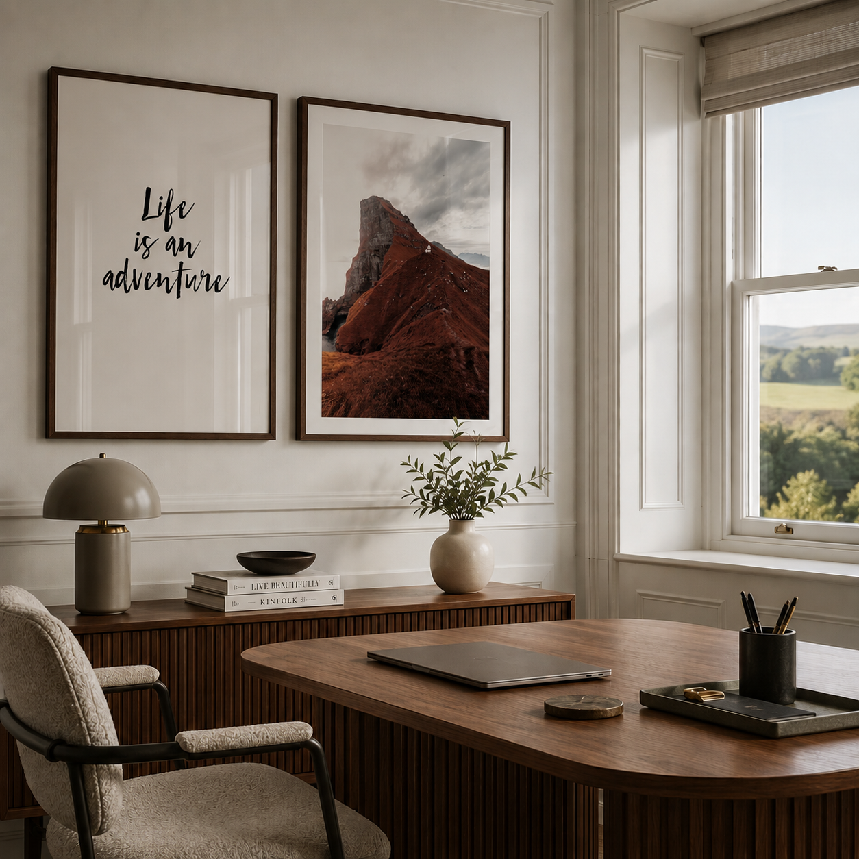 A styled home office interior with two framed wall art prints hanging above a wooden desk. The left print reads “Life is an adventure” in black script, and the right print shows a red rock mountain landscape.