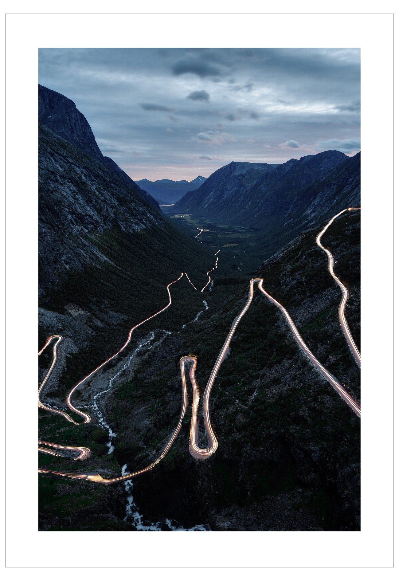Winding mountain road at night with lights on, surrounded by dark mountains and clouds.