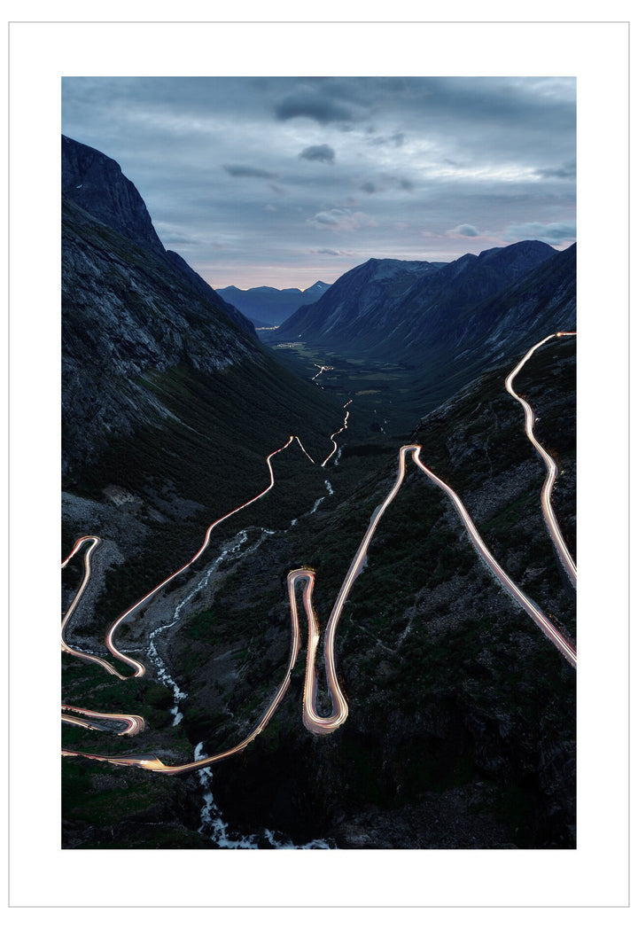 Winding mountain road at night with lights on, surrounded by dark mountains and clouds.