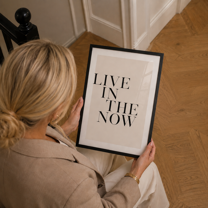 Person holding a framed quote 'LIVE IN THE NOW' on a wooden floor.